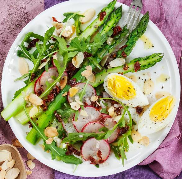 Salat aus grünem Spargel, Radieschen und Rucola auf einem Teller getoppt mit Ei, daneben Dressing