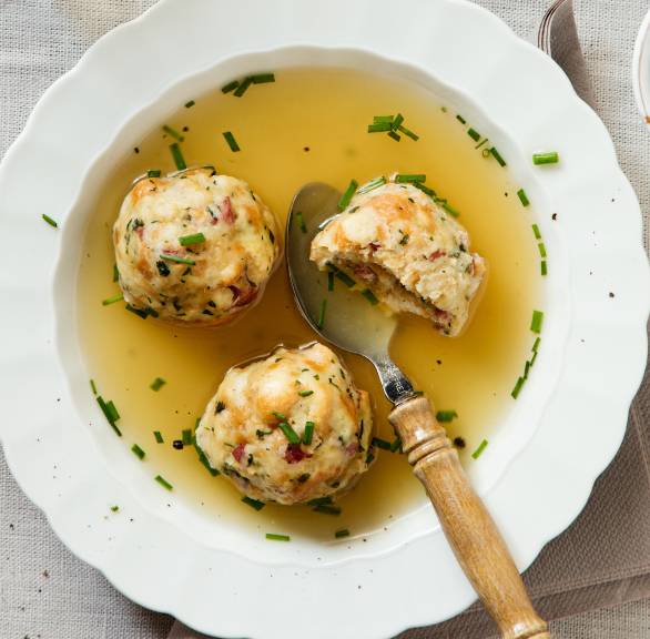 Südtiroler Speckknödel in einem Suppenteller mit Brühe, daneben frischer Schnittlauch auf einem Holzbrett