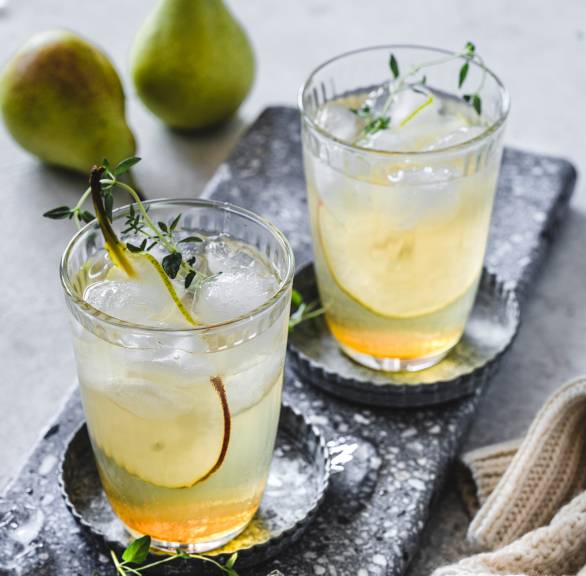 Birnen-Lillet mit Zimtsirup in zwei Gläsern mit Eiswürfeln garniert mit Thymian auf einem Tisch