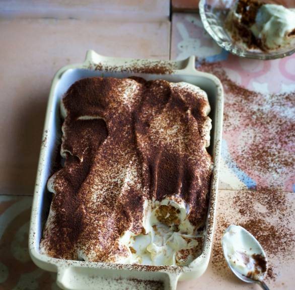 Tiramisu in einer Auflaufform mit einem kleinen fehlenden teil, daneben ein benutzter Löffel auf einem Tisch