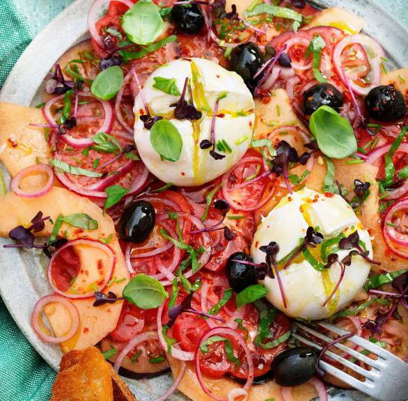 Eine Kugel Burrata auf einem Bett aus Melonen- und Tomatenscheiben getoppt von Zwiebeln und Oliven