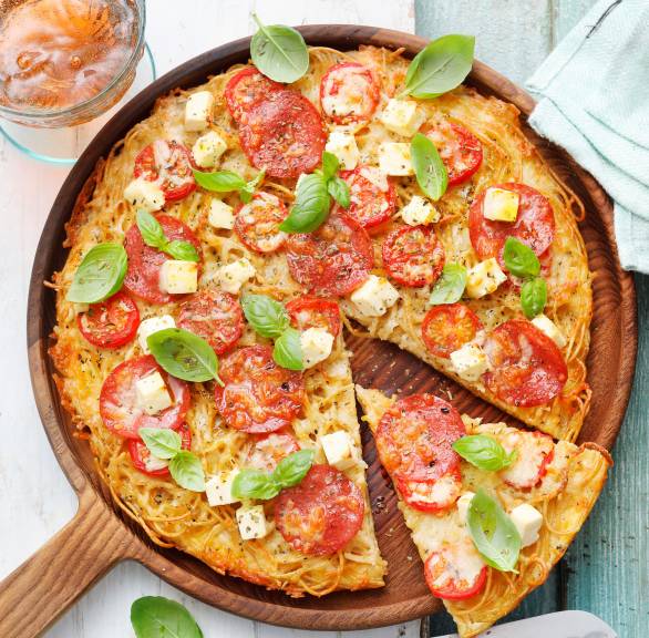 Runde Spaghettipizza mit Tomaten, Mozzarella und Feta angeschnitten auf einem Holzteller