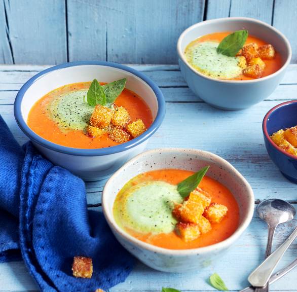 Tomatensuppe in mehreren Suppenschalen getoppt mit mit Basilikumschaum und Parmesan-Polenta-Croûtons auf einem Holztisch