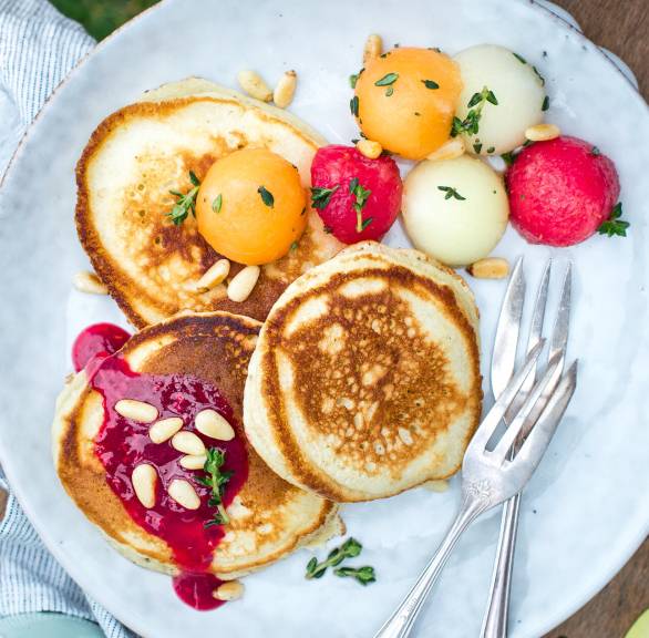 Pancakes auf einem Teller getoppt von Melonenkugeln und Himbeersauce auf einem Gartenstuhl