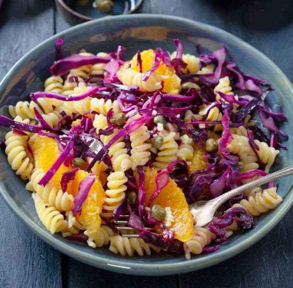 Nudelsalat mit Rotkohl, Kapern und Orangen angerichtet auf einem tiefen Teller, daneben Dressing in einer Schale