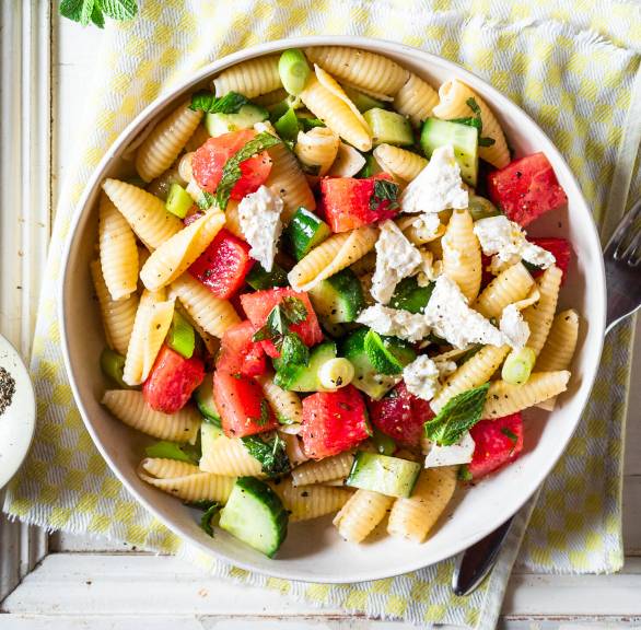 Pastasalat in einer Schüssel mit Wassermelonenstücken, Gurke, Fetawürfeln und Minzblättern auf einem Tisch
