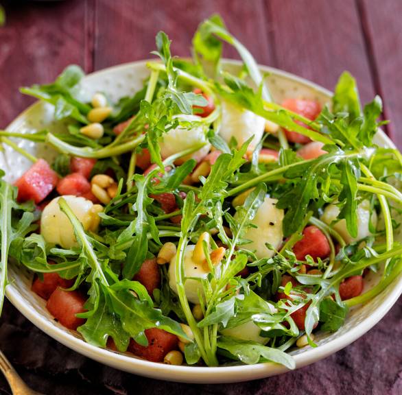 Wassermelonensalat mit Mini-Mozzarellakugeln und Rucola auf einem Teller, daneben Pinienkerne