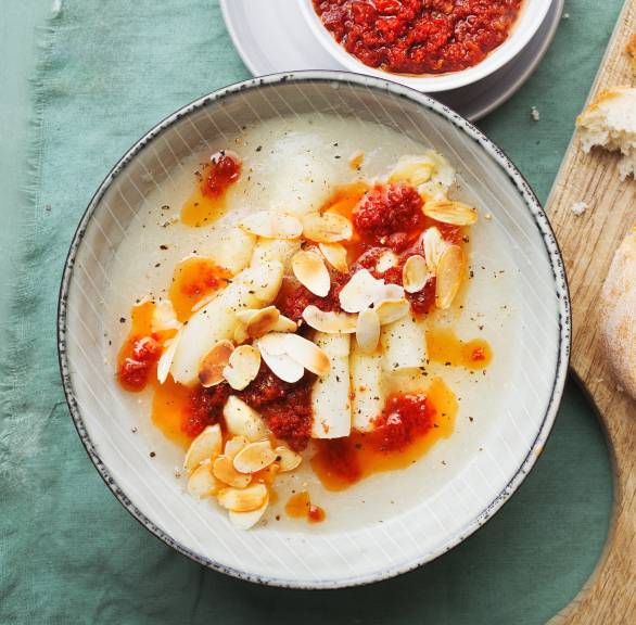 Weiße Spargelsuppe in einer Schale getoppt mit Tomatenöl und Mandelblättchen, daneben einige Scheiben Ciabatta