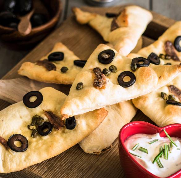 Pizzaecken mit Sardellen und Oliven auf einem Holzbrett, daneben Rosmarindip