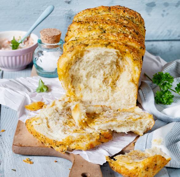 Zupfbrot auf einem Holzbrett, es fehlen einige Scheiben, daneben Salz und Knoblauch auf einem Tisch