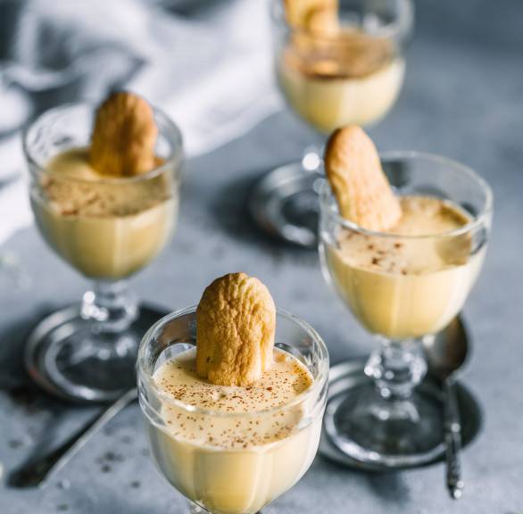 Zabaione in vier Gläsern getoppt von jeweils einem Löffelbiskuit und Marsala auf einem Tisch