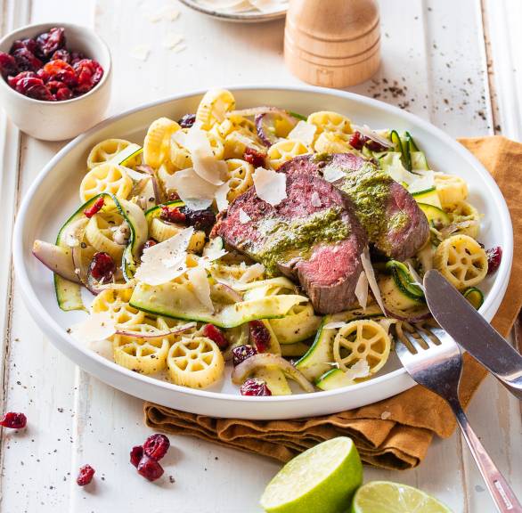 Italienischer Nudelsalat mit Zucchini angerichtet mit gebackenen Rinderfiletscheiben und grünem Pesto-Dressing auf einem Teller aus dem Backofen