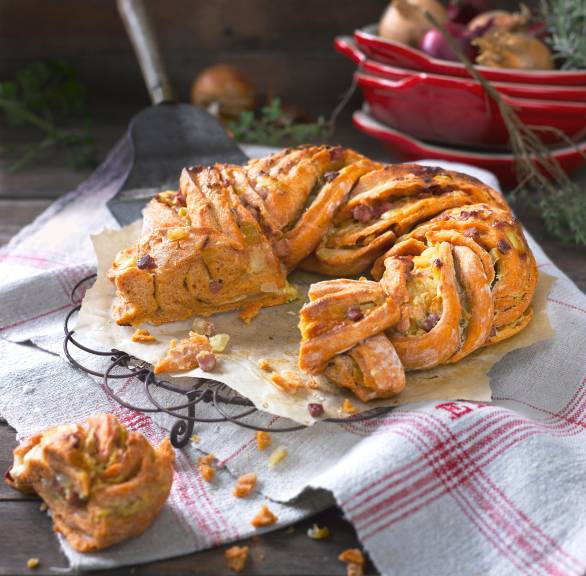 Gefüllter Brotkranz angeschnitten auf einem Tisch, dahinter Zwiebeln in einer Schale