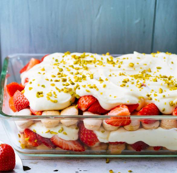 Erdbeertiramisu mit Quark und Joghurt in einer gläsernen Auflaufform bestreut mit Pistazien, daneben frische Erdbeeren