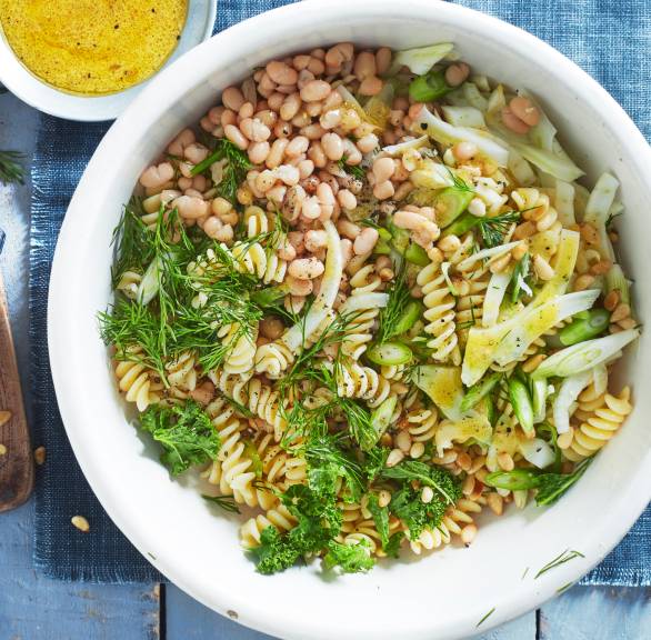 Veganer italienischer Nudelsalat mit Fenchel, Grünkohl und Bohnen in einer Salatschüssel, daneben Pinienkerne und Dressing