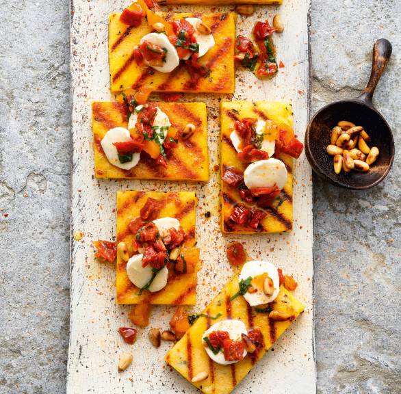 Polentaschnitten in Rechtecke geschnitten mit Grillstreifen getoppt von Tomaten, Mozzarella und Pinienkernen auf einem Teller