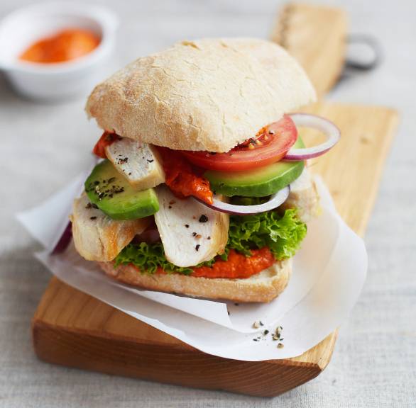 Ein Ciabatta-Burger belegt mit Hähnchenbrust, Avocado und pikanter Ajvar-Creme angerichtet auf einem Holzbrett
