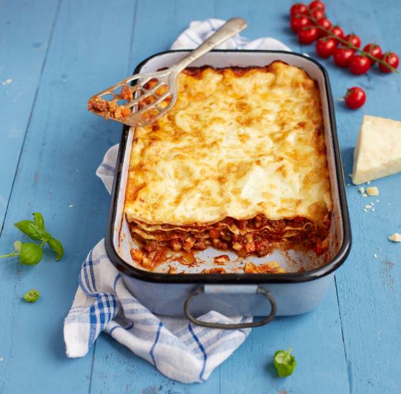 Lasagne mit Bolognese-Schichten in einer Auflaufform angeschnitten auf einem Tisch, daneben Parmesan und Tomaten
