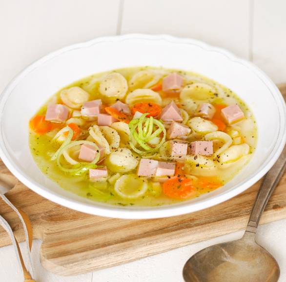 Nudelsuppe in einem Teller auf einem Holzbrett, daneben Mortadellastücke