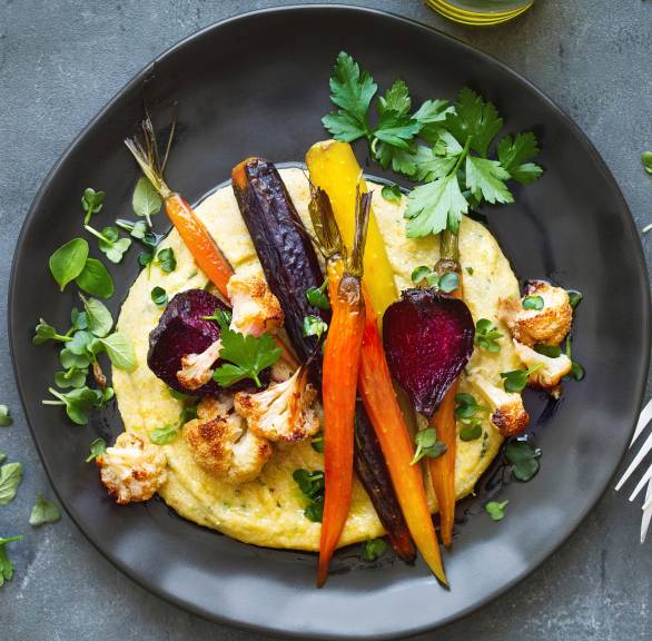 Cremige Polenta auf einem Teller getoppt von Gemüse aus dem Ofen, darauf frische Kräuter