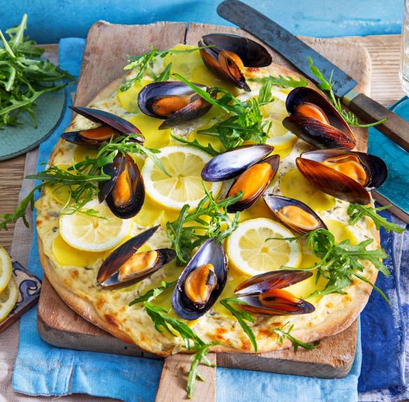 Ovale Pizza Bianca mit Miesmuscheln, Kartoffeln und Rucola auf einem Holzbrett garniert mit Zitronenscheiben