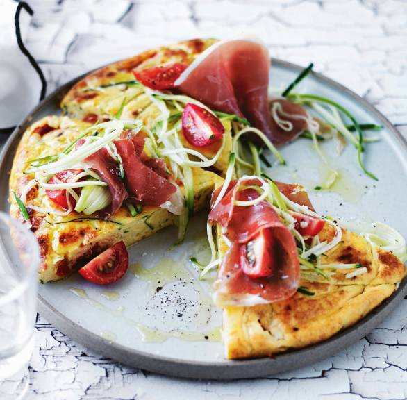 Runde Zucchini-Frittata aus Polenta angeschnitten auf einem Teller getoppt von Kirschtomaten und rohem Schinken