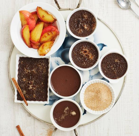 Fünf Ofenförmchen mit Schokoladen-Crème-Brûlée auf einem Tablett, daneben eine Schale mit karamellisierten Äpfeln