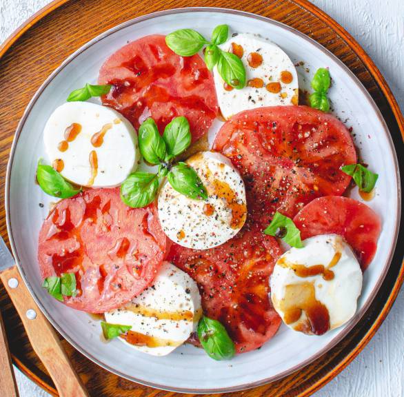 Tomaten- und Mozzarellascheiben auf einem Holzteller angerichtet als Caprese-Salat getoppt mit Balsamicodressing