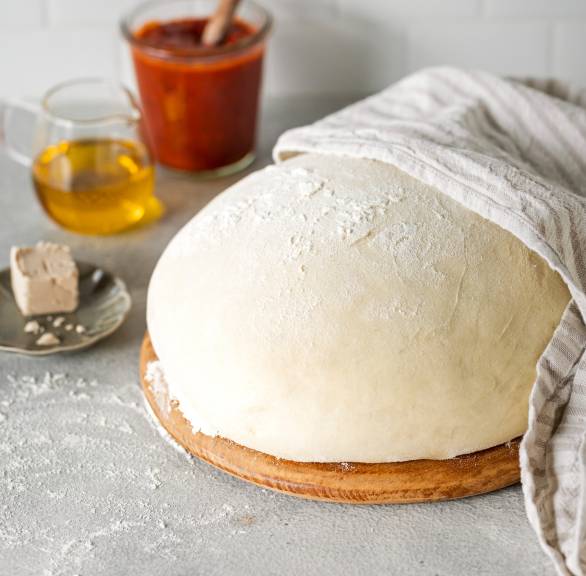 Pizzateig als Kugel geformt und bemehlt auf einem Holzbrett, daneben Olivenöl und frische Hefe