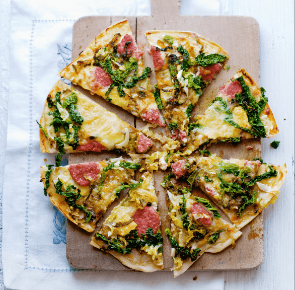 Runder Flammkuchen mit Wirsing, Schmand und Mettwurst Belag in Pizzastücke geschnitten auf einem Holzbrett