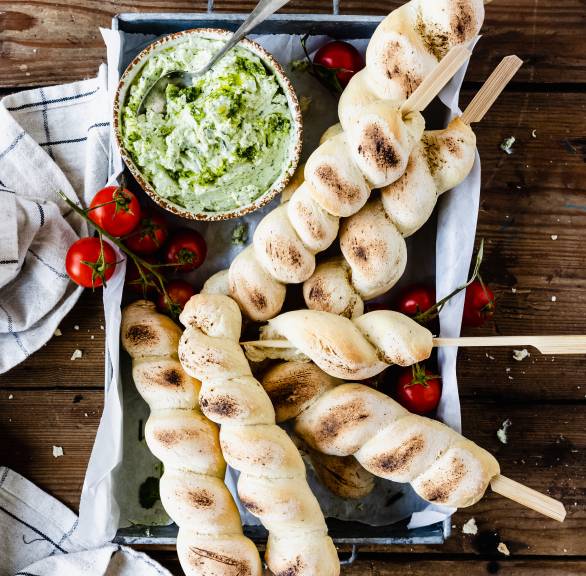Fünf gebackene Stockbrot auf einem Backblech auf einem Holztisch, daneben Dip und Strauchtomaten