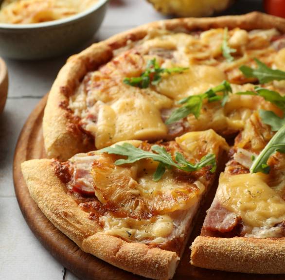 Runde Pizza Hawaii angeschnitten auf einem Holzbrett, dahinter eine halbierte Ananas