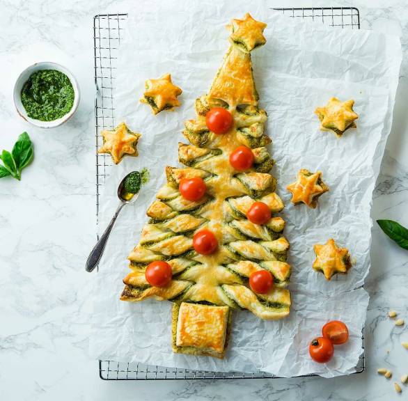 Tannenbaum aus Blätterteig auf einem Kuchengitter, daneben Kirschtomaten, grünes Pesto und Basilikum