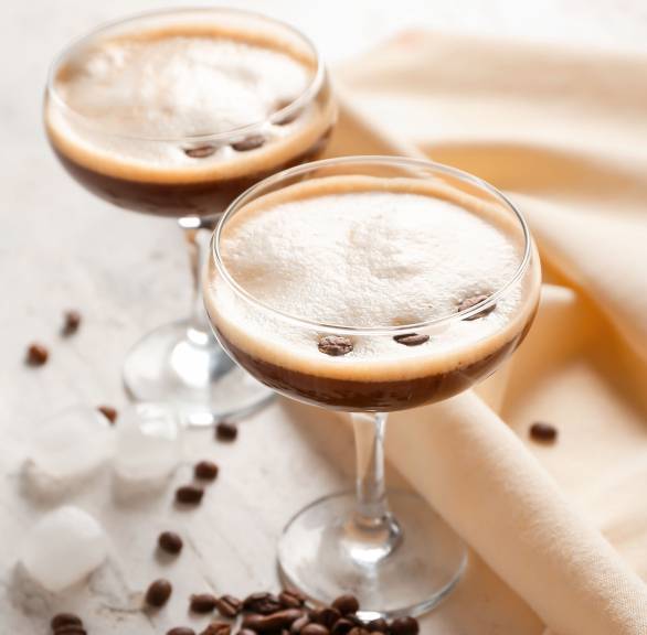 Espresso Martini in zwei Champagnergläsern auf einem Tisch, daneben Kaffeebohnen und Eiswürfel