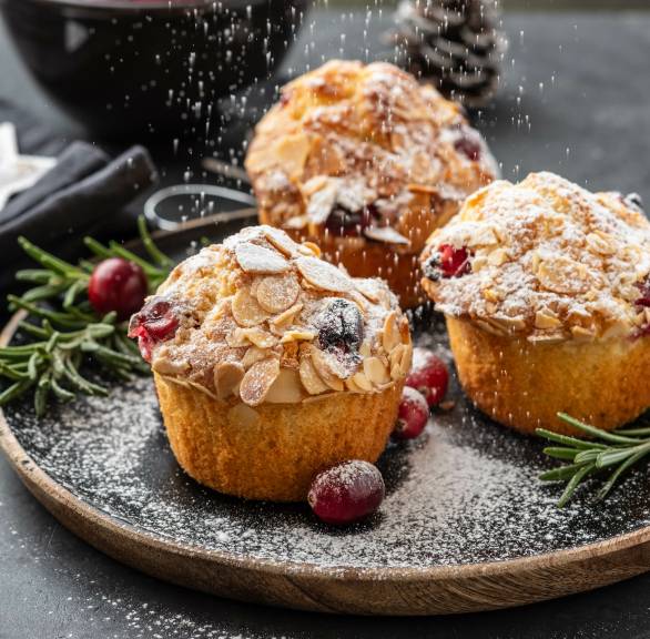 Weihnachtliche Muffins mit Cranberry, Rosmarin & Mandeln auf einem schwarzen Teller werden gerade mit Puderzucker bestäubt