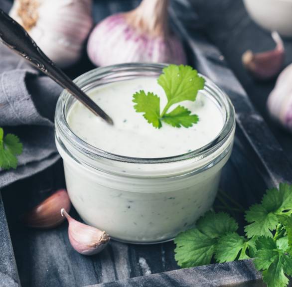 Joghurtdip in einem Glasbescher, daneben Knoblauch, Olivenöl, Minze und Zitrone