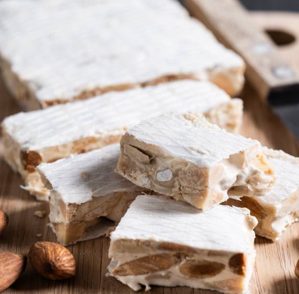 Rechteckiger Torrone angeschnitten auf einem Holzbrett, daneben ganze Mandeln