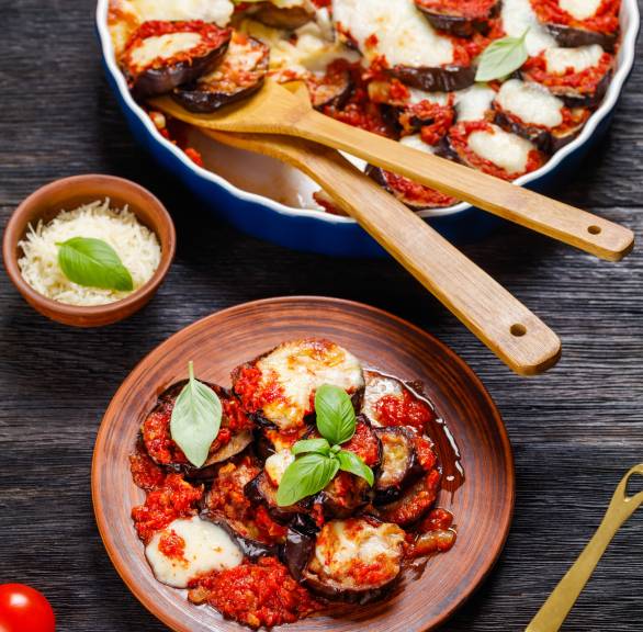 Veganer Auberginen-Auflauf Parmigiana in einer runden Auflaufform, daneben eine Portion auf einem Teller sowie frischer Basilkum