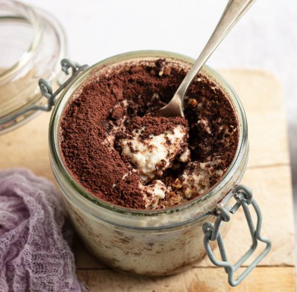 Veganes Tiramisu in einem geöffneten Weckglas auf einem Holzbrett, darin ein Dessertlöffel