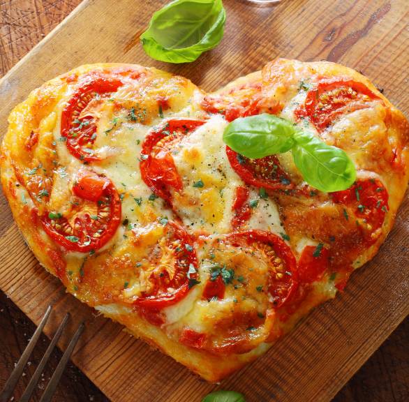 Pizza in Herzform auf einem Holztisch garniert mit Oregano und Basilikum, daneben ein Glas Wein