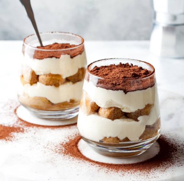Zwei Gläser mit Tiramisu auf einer Marmorplatte bestäubt mit Kakaopulver, im Hintergrund eine Espressokanne