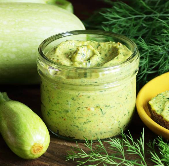 Selbst gemachtes Zucchinipesto in einem Einmachglas auf einem Holztisch, daneben Zucchinis und Kräuter