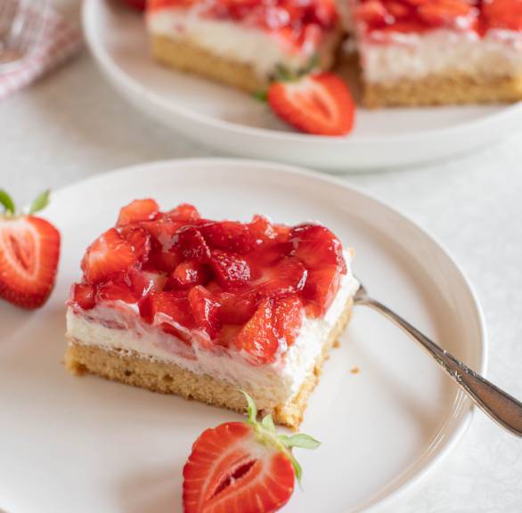 Ein Stück Erdbeerkuchen mit Pudding-Quark-Creme, dahinter das Blech mit dem restlichen Blechkuchen