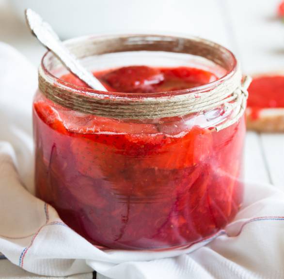Selbst gemachte Erdbeermarmelade in einem Einmachglas, dahinter frische Erdbeeren auf einem Holztisch