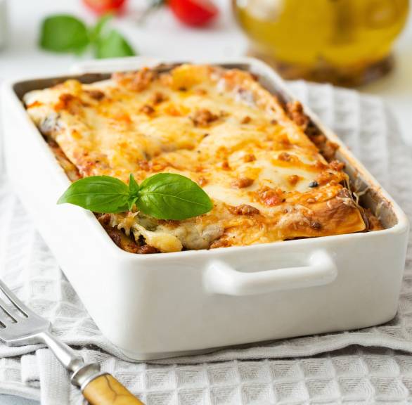 Gemüselasagne in einer Auflaufform auf einem Tisch, dahinter Olivenöl und Tomaten