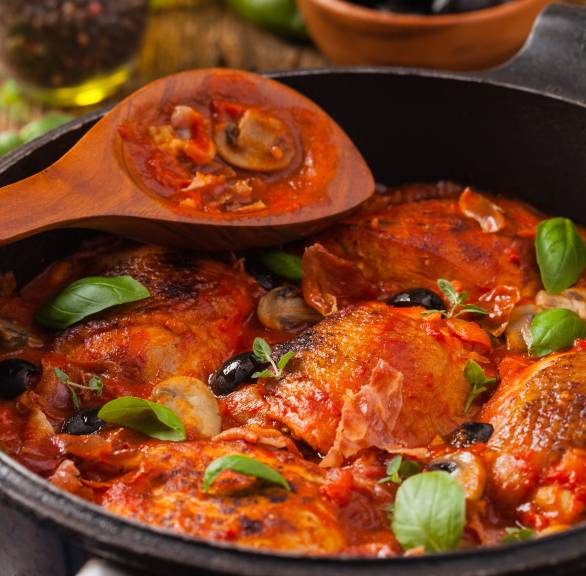 Geschmorte Hähnchenschenkel alla cacciatora in Tomatensauce in einer Pfanne garniert mit schwarzen Oliven und frischen Kräutern