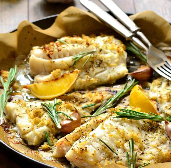 Kabeljau aus dem Ofen in einer Auflaufform garniert mit Zitronenspalten und Rosmarinzweigen auf einem Holztisch