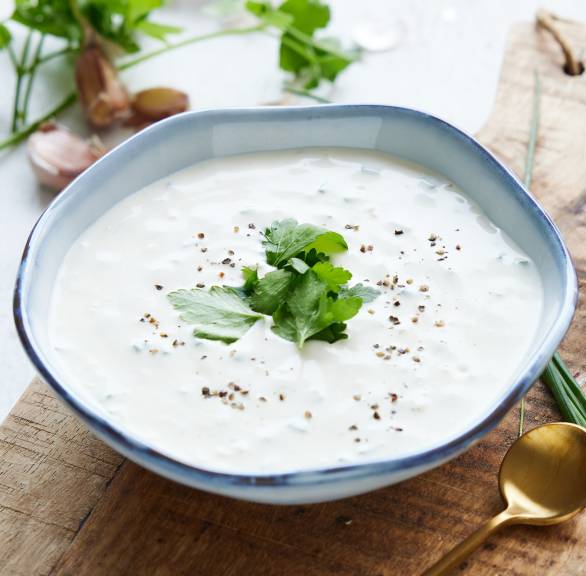 Knoblauch-Dip in einer wellenförmigen Schale auf einem Holzbrett garniert mit frischen Kräutern, daneben Knoblauchzehen