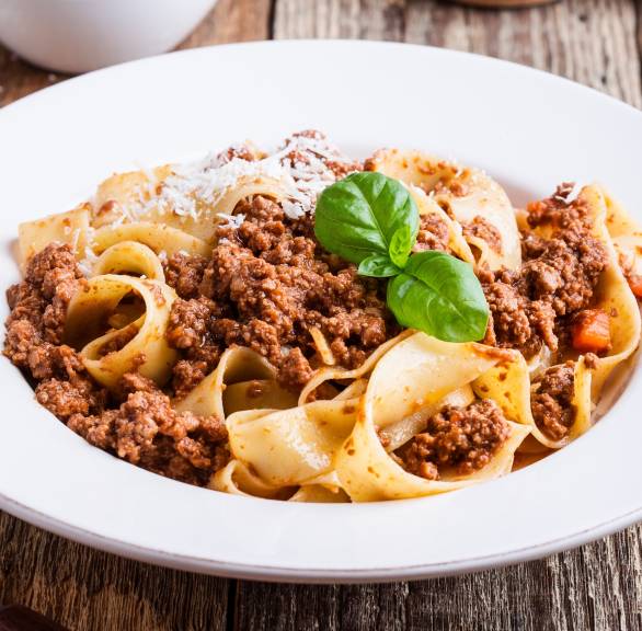 Pappardelle mit Bolognese aus Kalbshackfleisch auf einem tiefen weißen Teller, daneben Parmesan und Gewürze auf einem Holztisch