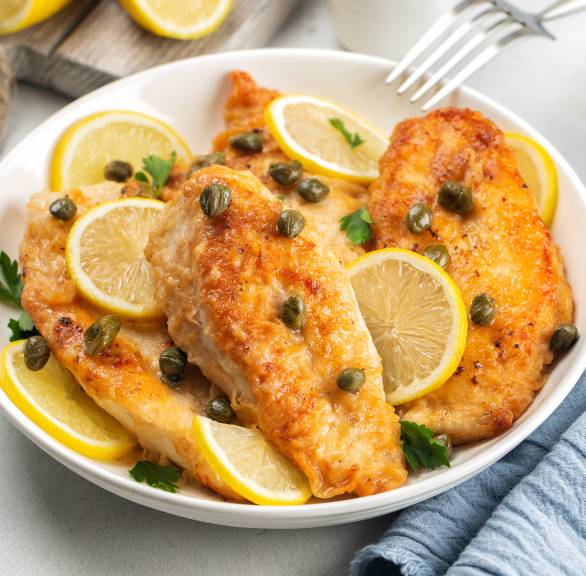 Putenbruststücke als Piccata alla milanese auf einem Teller übergossen mit Zitronen-Kapern-Sauce dekoriert mit Zitronenscheiben und frischer Petersilie